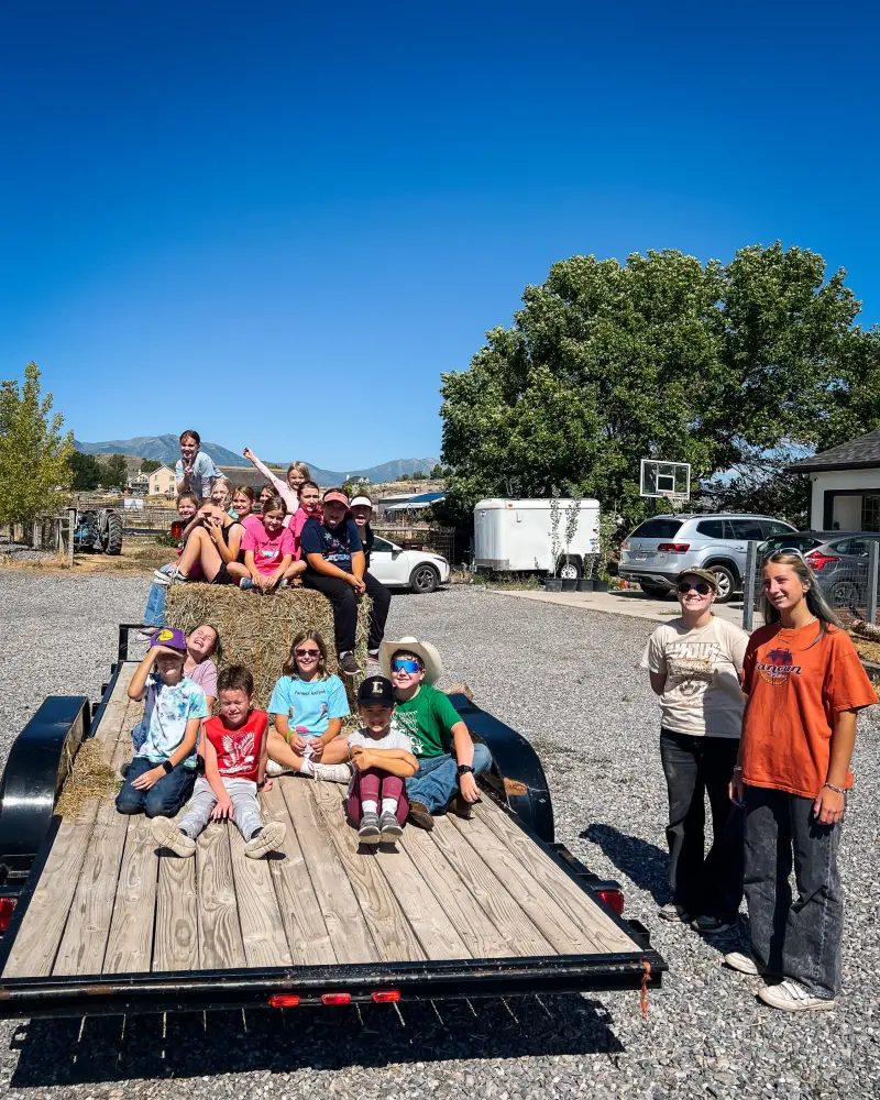 Campers waving from a hay bale on a flatbed trailer under a blue sky