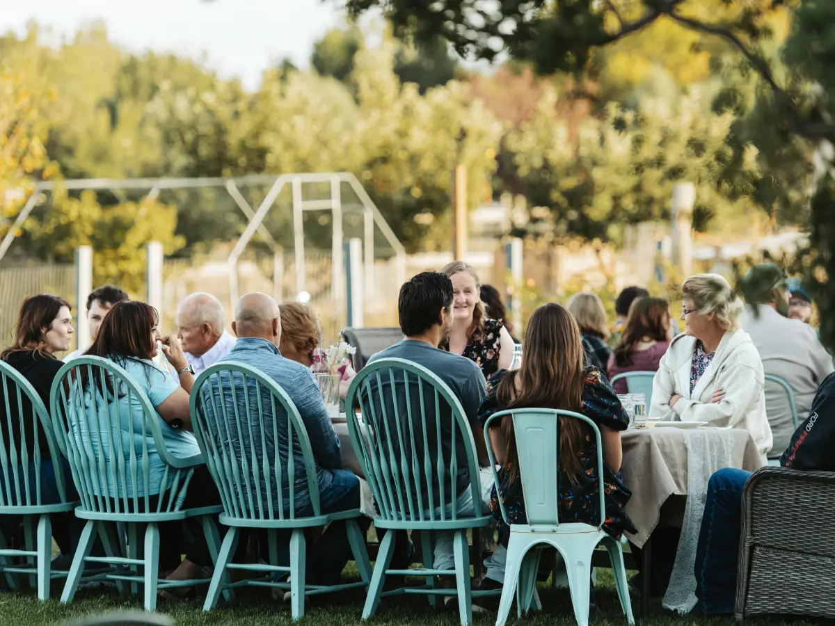 Guests talking over dinner at a long table on the lawn, seated on mint-green chairs under the trees