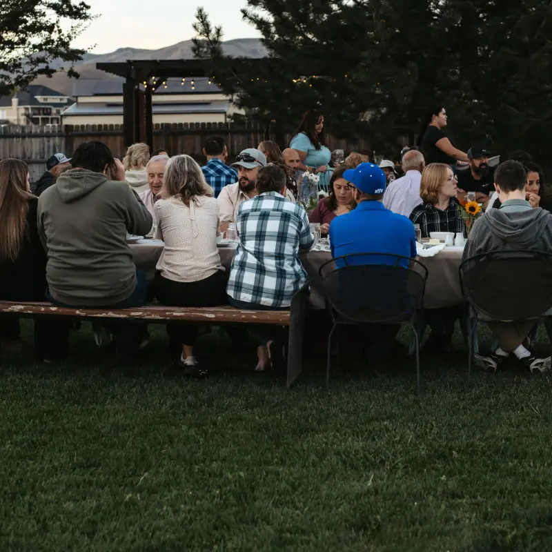 The full dinner table of guests on the lawn as evening falls, the pergola and mountains behind